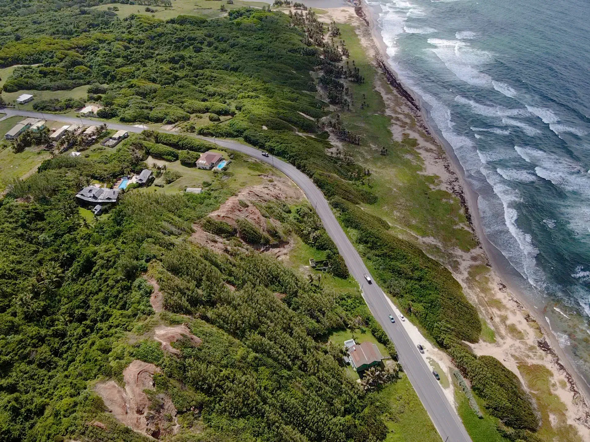 Aerial view of sloping coastal land, homes and a winding road beside the beach on Barbados’ east coast.